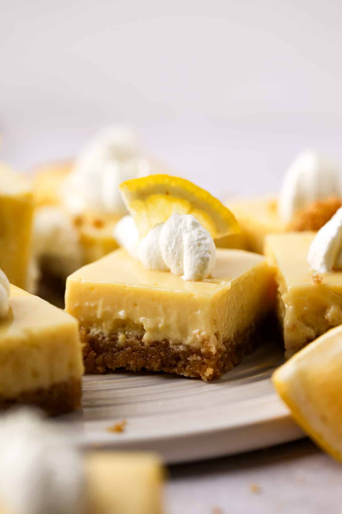 Close of up cut lemon bars with graham cracker crust topped with a piping of whipped cream and a slice of lemon.
