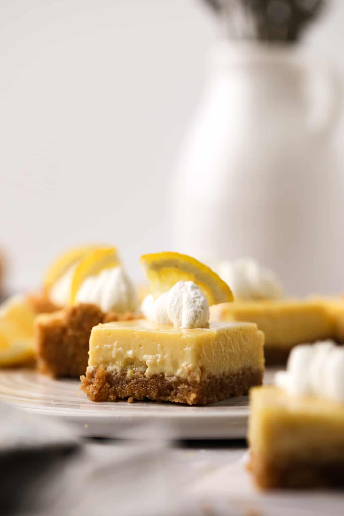 Close of up cut lemon bars with graham cracker crust topped with a piping of whipped cream and a slice of lemon.