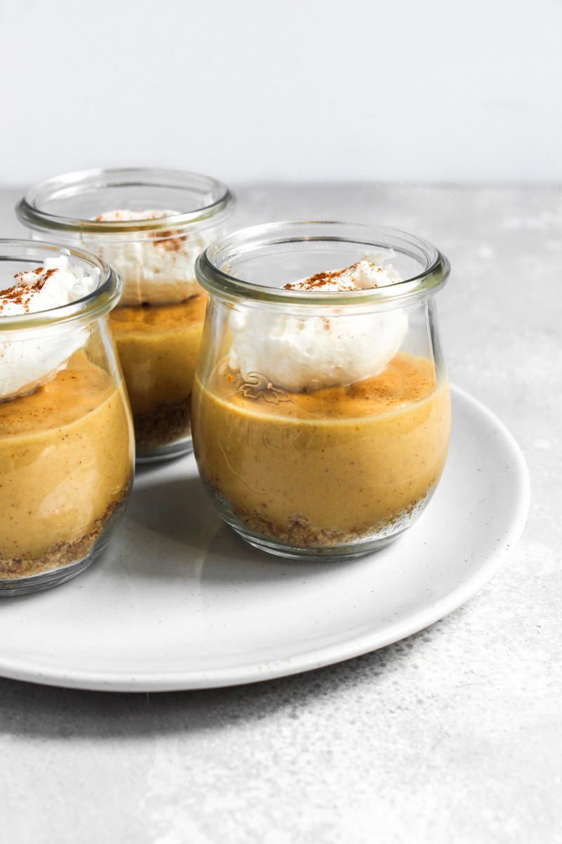 Jars of pumpkin custard with layers of graham cracker, custard, and whipped cream on a plate.