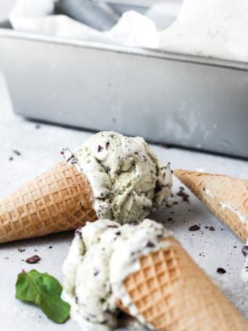 Three mint chocolate chip ice creams with sugar cones on their sides next to container of ice cream.