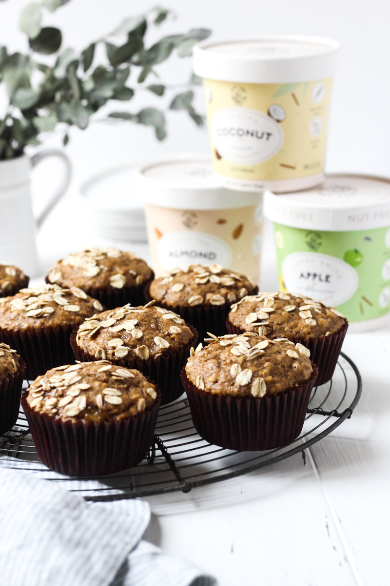 Baked banana muffins on a round french iron cooling rack with stack of mylk labs oatmeal cups in the background.
