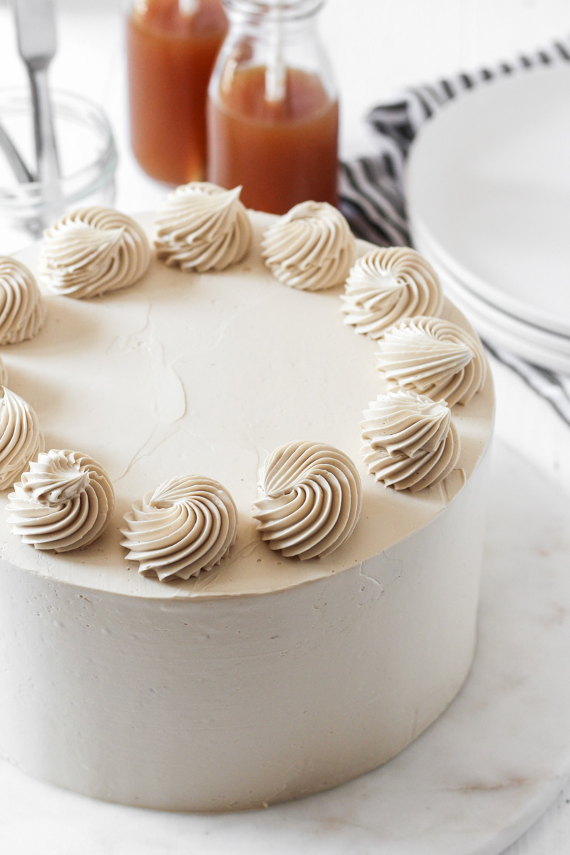 Looking over an 8-inch round apple cider cake with brown sugar Swiss meringue buttercream with rosettes on top and apple cider in the background.