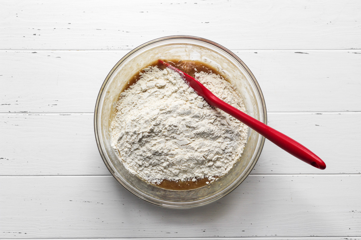 Banana bread cookie dough wet ingredients topped with dry ingredients in a glass bowl with a red spatula.
