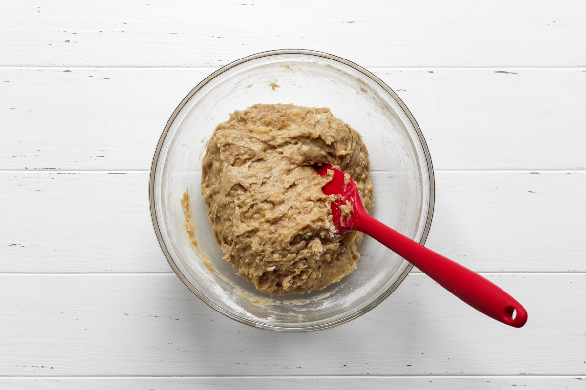 Banana bread cookie dough in a glass bowl with a red spatula.