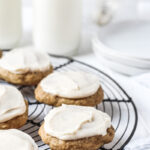 Banana bread cookies topped with cream cheese frosting on a French round wire rack with milk and plates in the background.