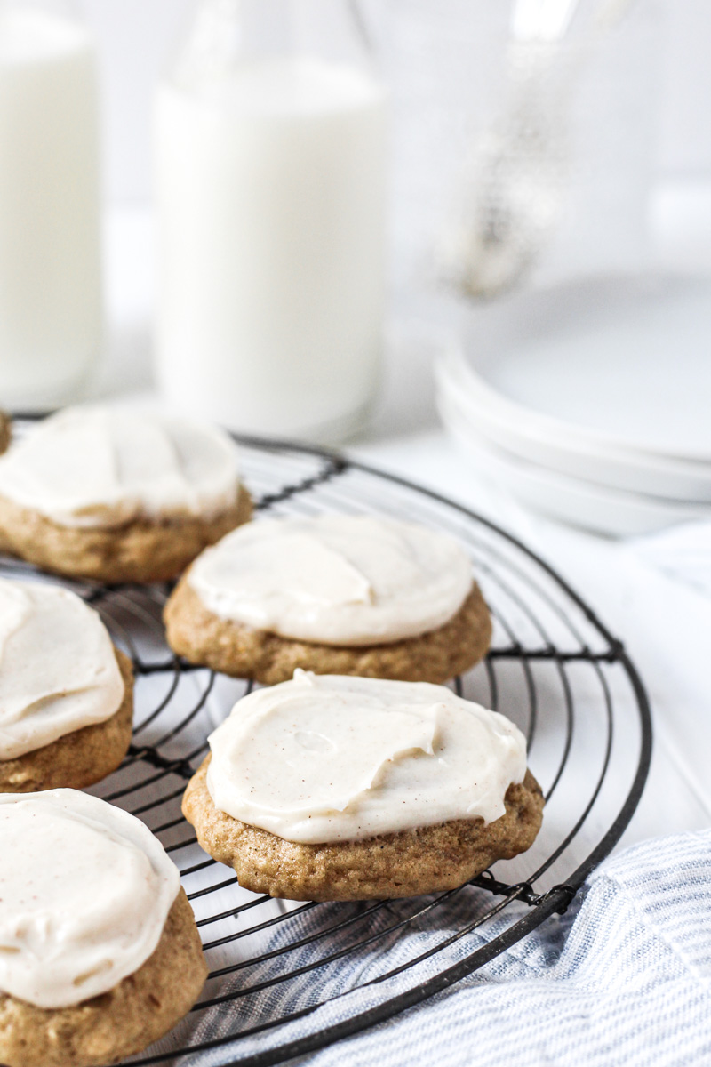 Banana bread cookies topped with cream cheese frosting on a French round wire rack with milk and plates in the background.