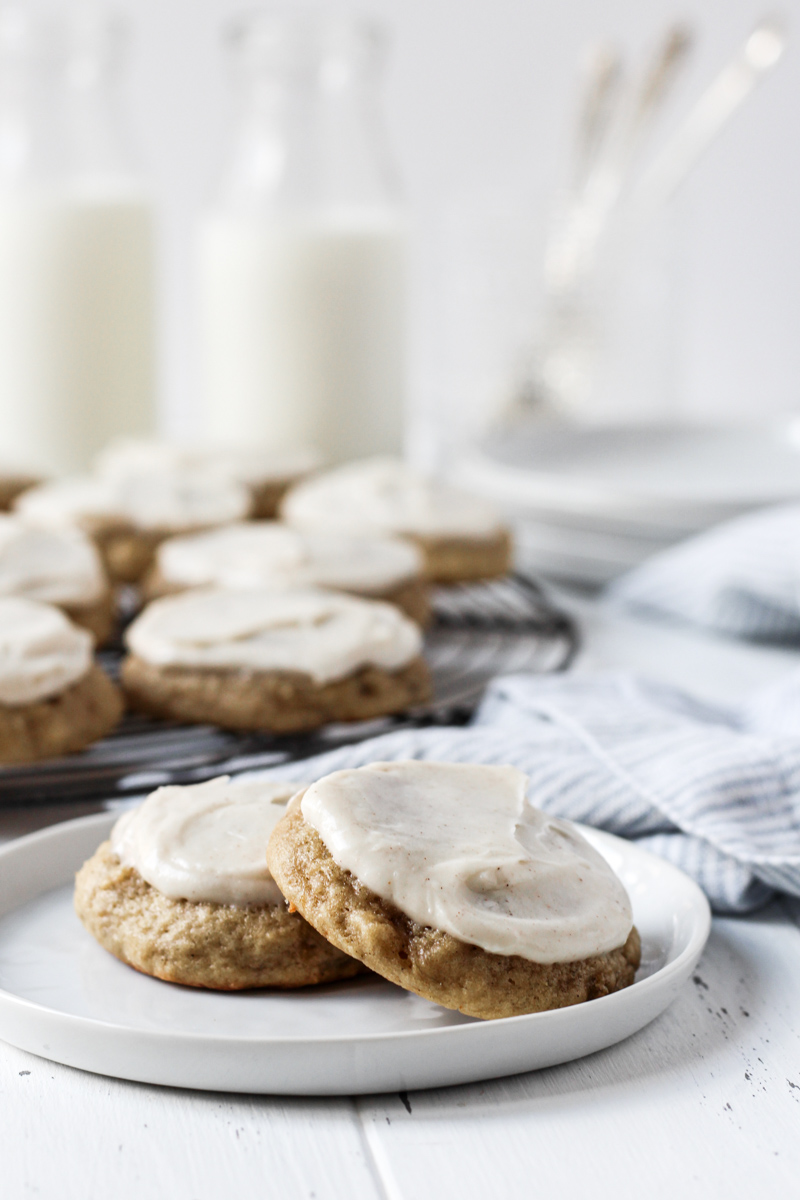 Two banana bread cookies topped with cream cheese frosting on a plate with more cookies on a French round wire rack, milk, and plates in the background.