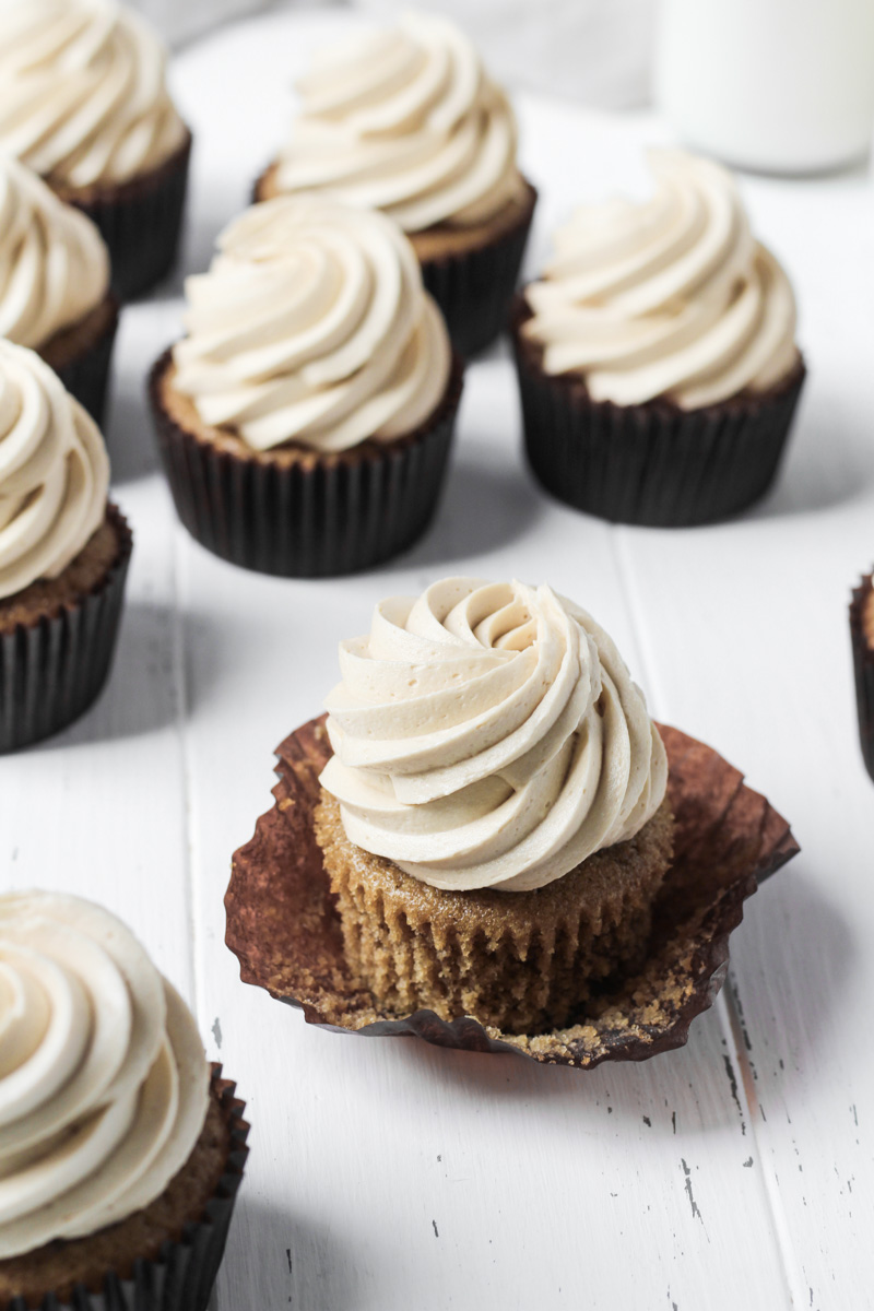 Several coffee cupcakes with coffee buttercream on a white wood board for display with one cupcake with the wrapper loosened.