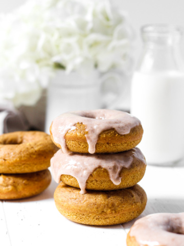 Stack of three baked pumpkin donuts, two with glaze and more in the background with flowers and milk.
