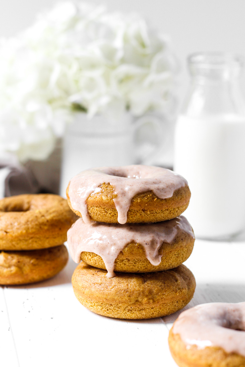 Stack of three baked pumpkin donuts, two with glaze and more in the background with flowers and milk.