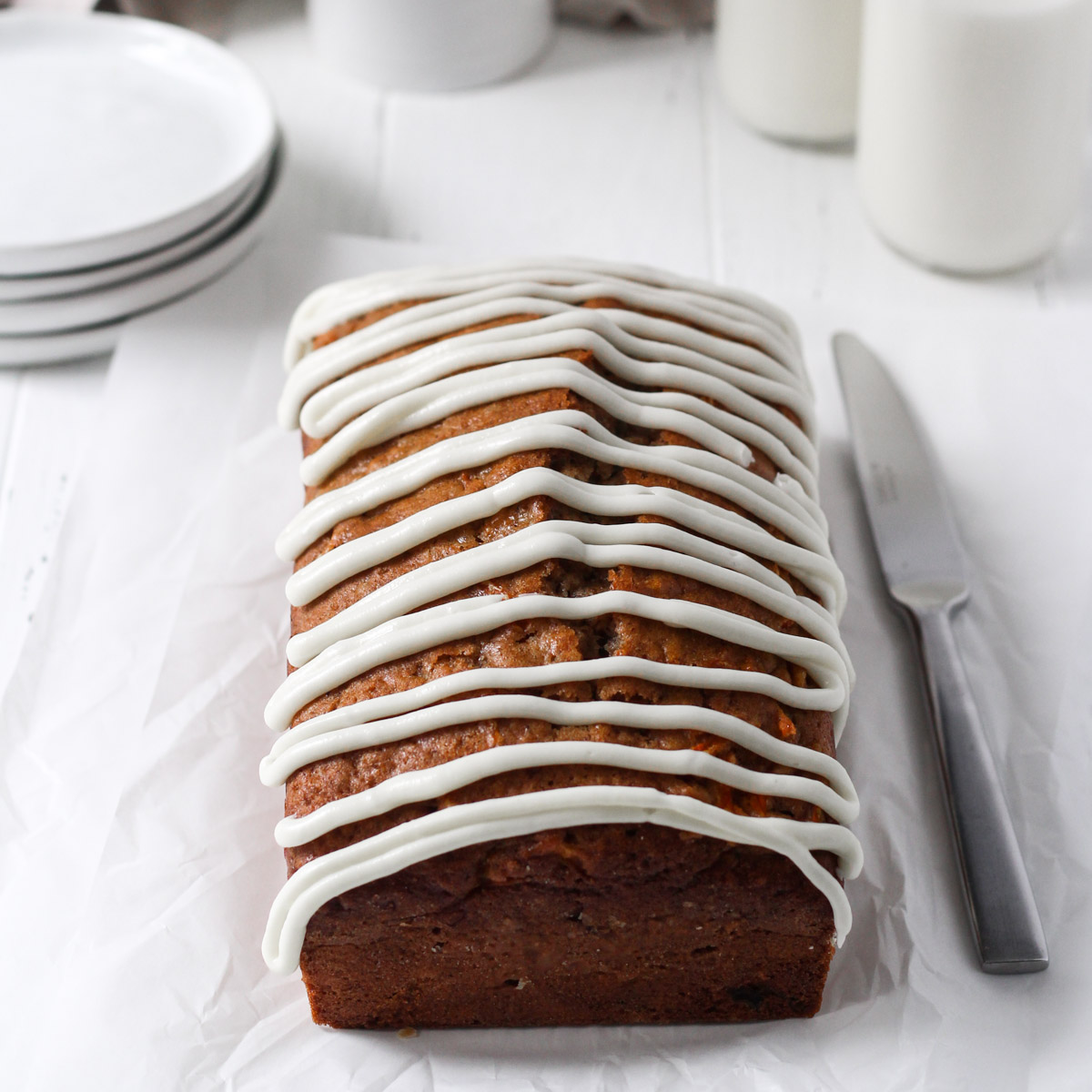 Carrot cake loaf with a zig zag drizzle of homemade cream cheese frosting on a piece of white parchment paper with two glasses of milk in the background.