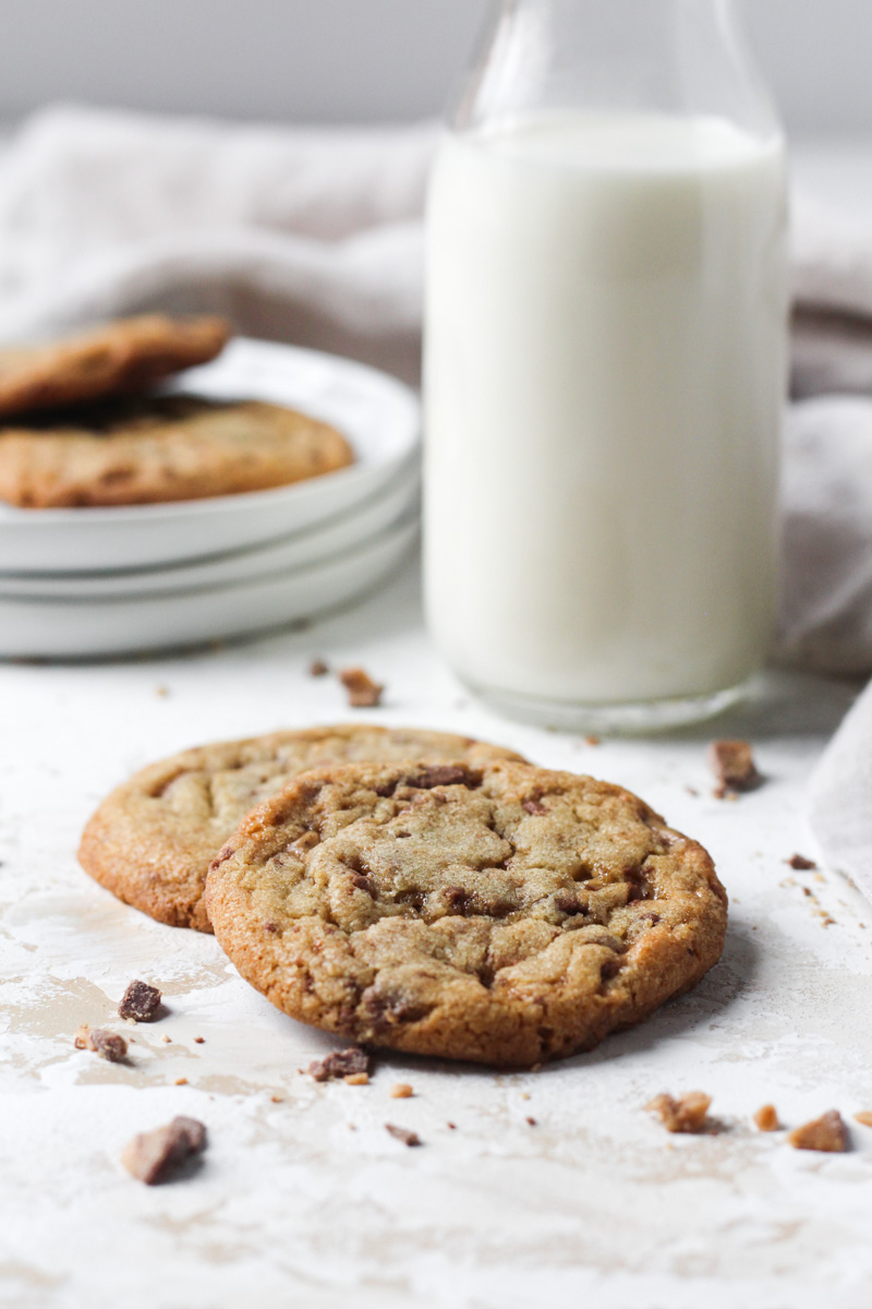 Close up of two brown butter cookies with toffee bits and a glass of milk and plate of cookies in the background.