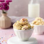 Small bowl of scoops of edible sugar dough mixed with sprinkles and with milk and flowers in the background.