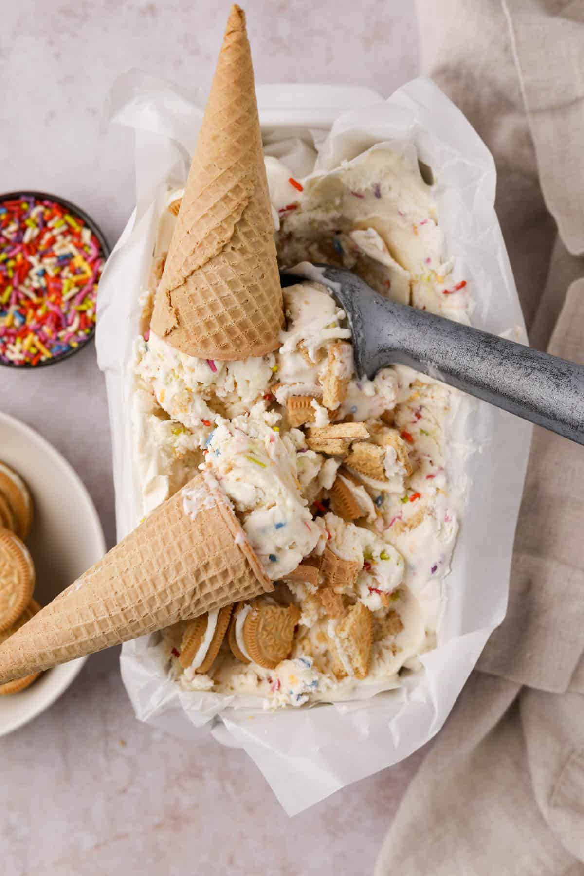 Frozen no churn Funfetti ice cream in a parchment lined loaf pan with an ice cream scoop, ice cream cones, Golden Oreos and sprinkles.