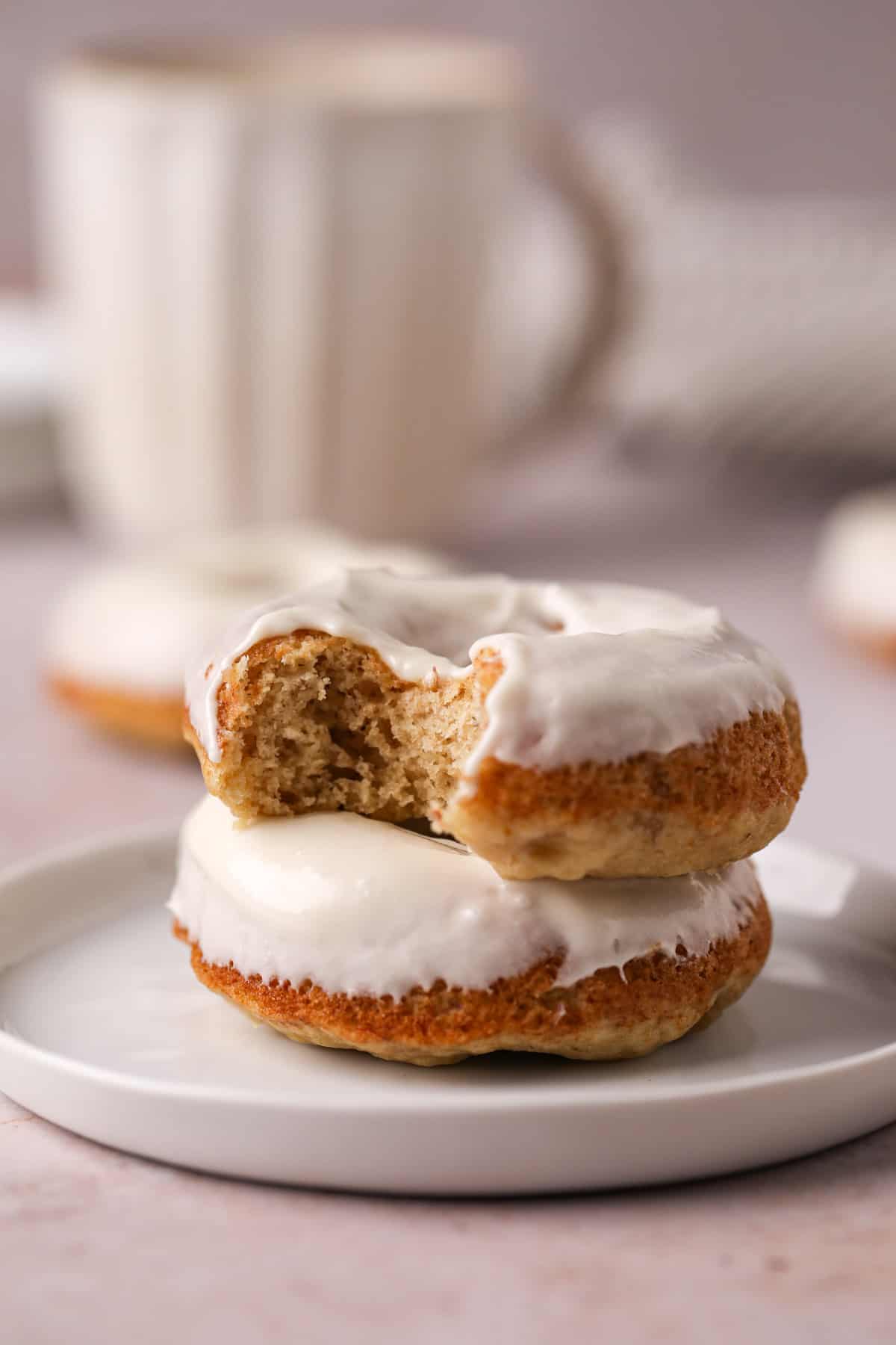 Stack of two cream cheese glazed baked banana donuts on a white plate with the top donut missing a bite of donut.