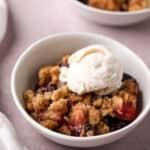 Baked cherry apple crumble in a white bowl topped with a scoop of vanilla ice cream.