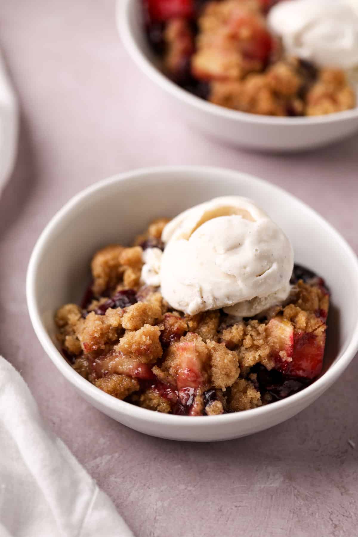 Baked cherry apple crumble in a white bowl topped with a scoop of vanilla ice cream.