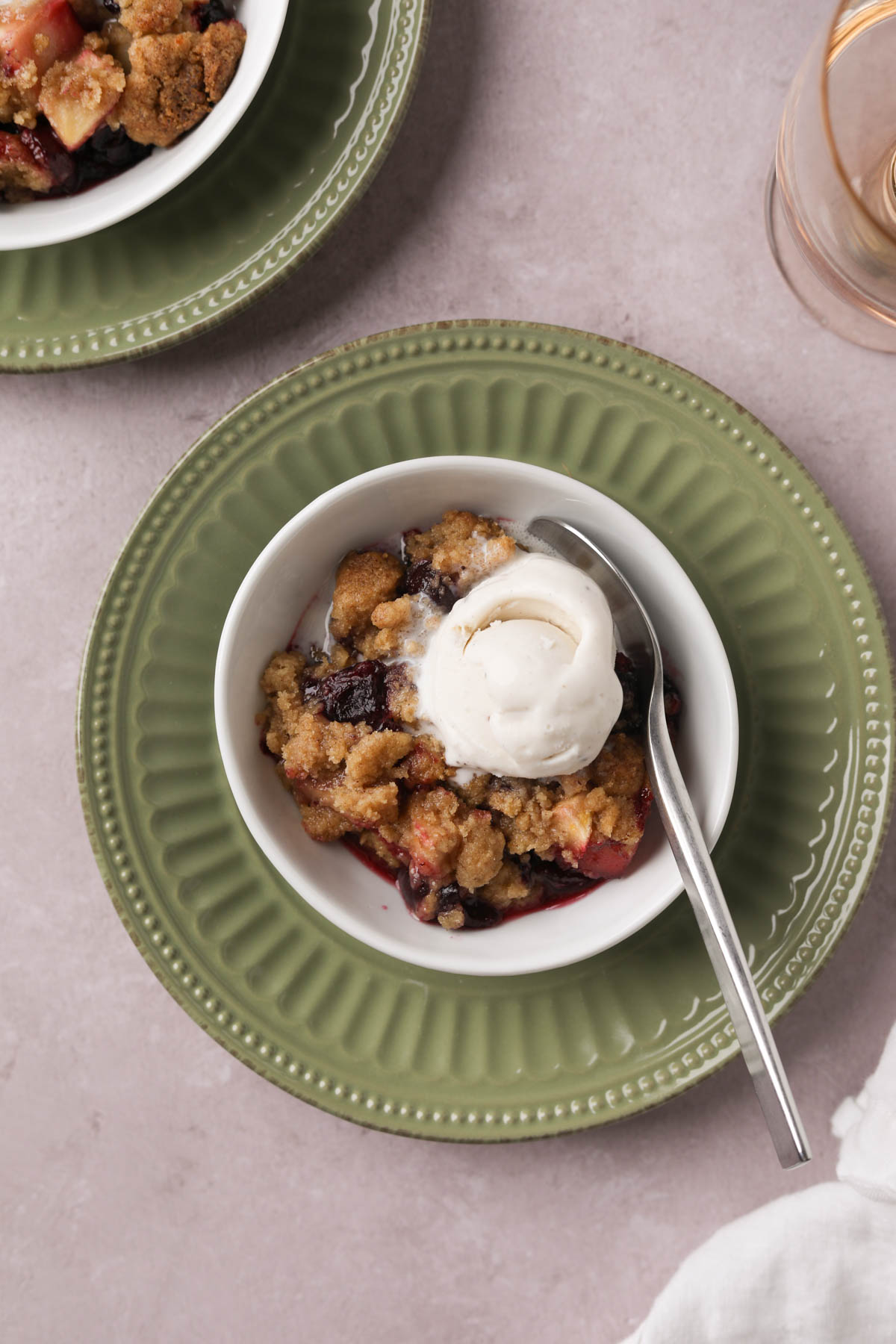 Overhead view of baked cherry apple crumble in a white bowl topped with a scoop of vanilla ice cream and spoon.