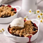 Individual peach cherry crisps baked in white handled ramekins with an oat crumble topping and a scoop of vanilla ice cream.