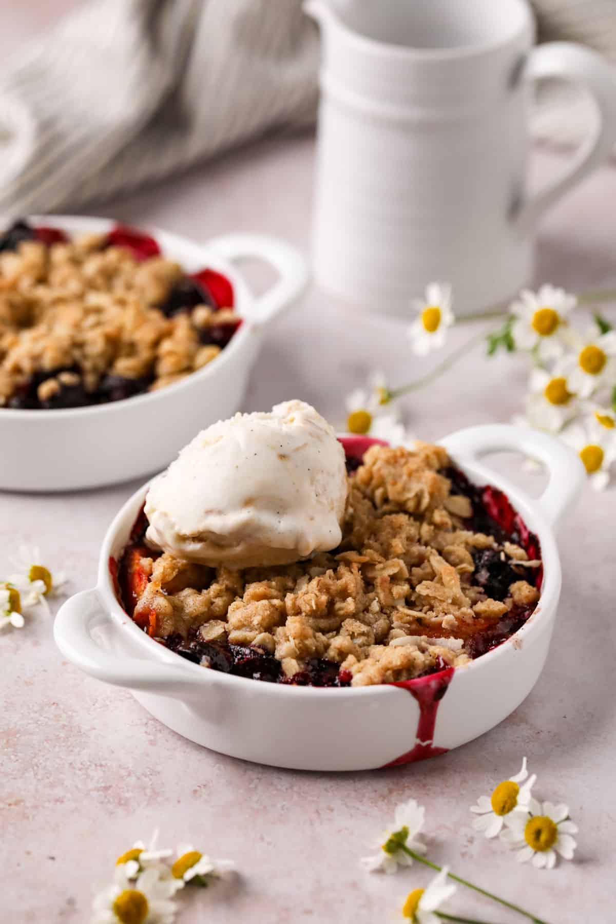 Individual peach cherry crisps baked in white handled ramekins with an oat crumble topping and a scoop of vanilla ice cream.