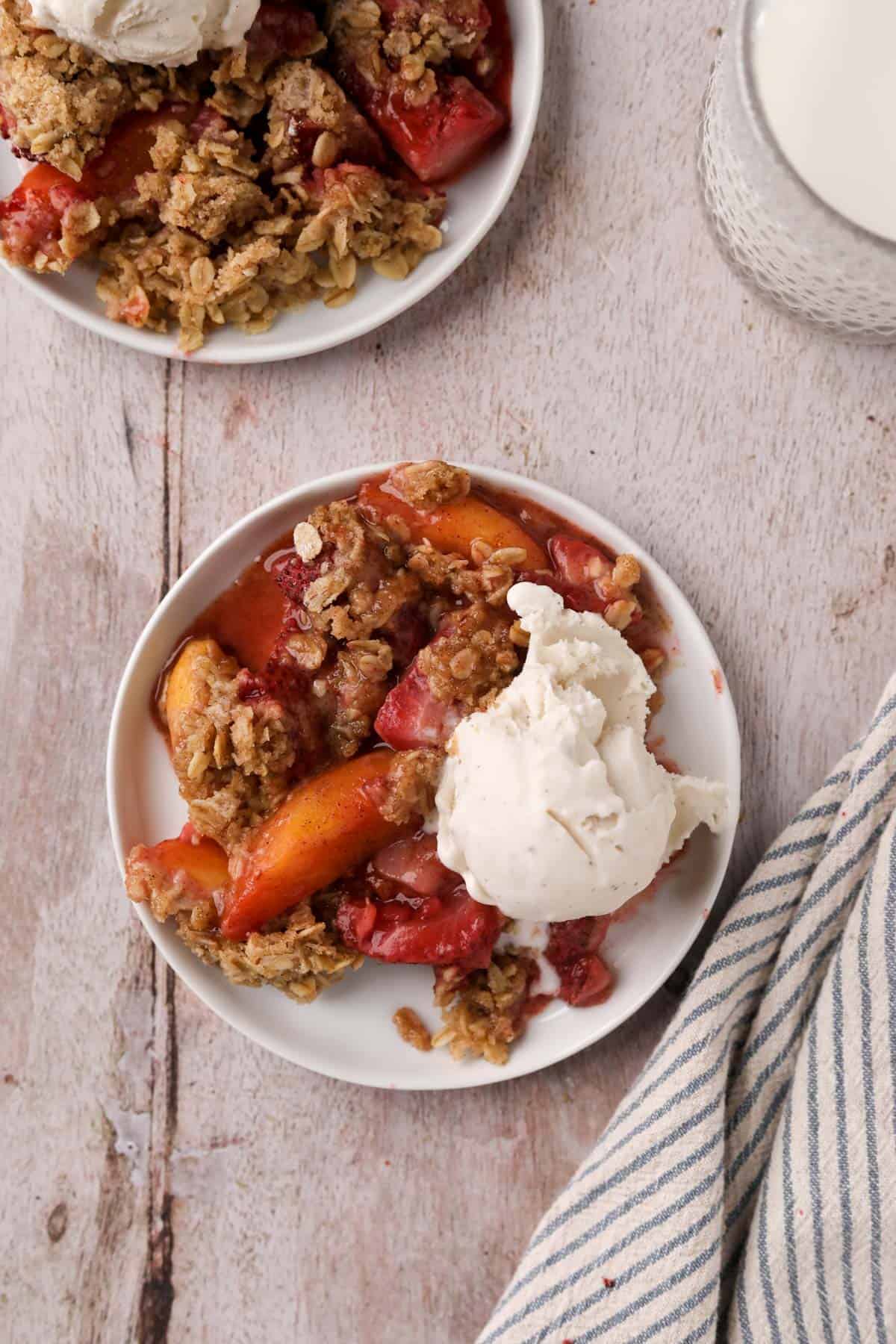 Overhead shot of two white dishes with strawberry peach crisp and a scoop of vanilla ice cream.