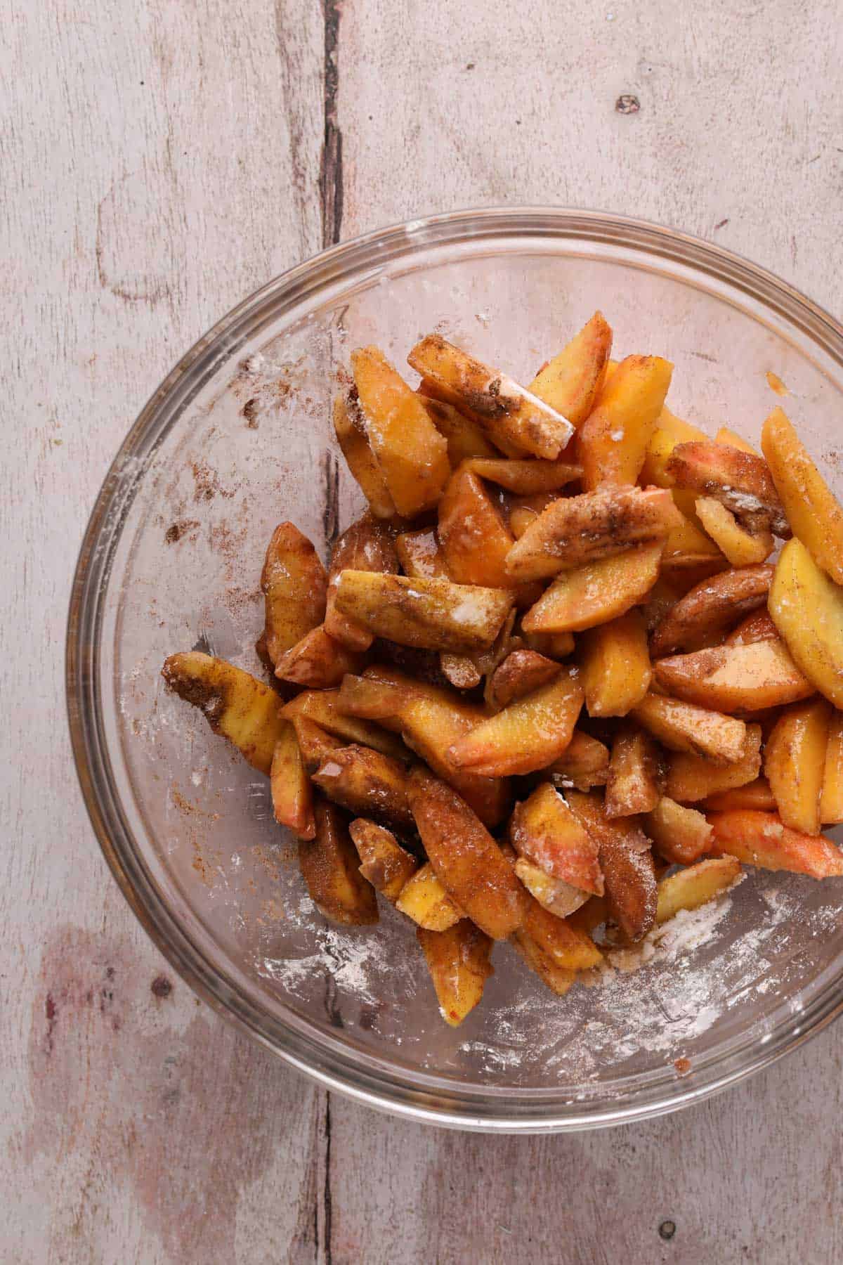 Sliced peaches mixed with sugar, cinnamon, cornstarch, lemon juice and vanilla in a large glass mixing bowl.