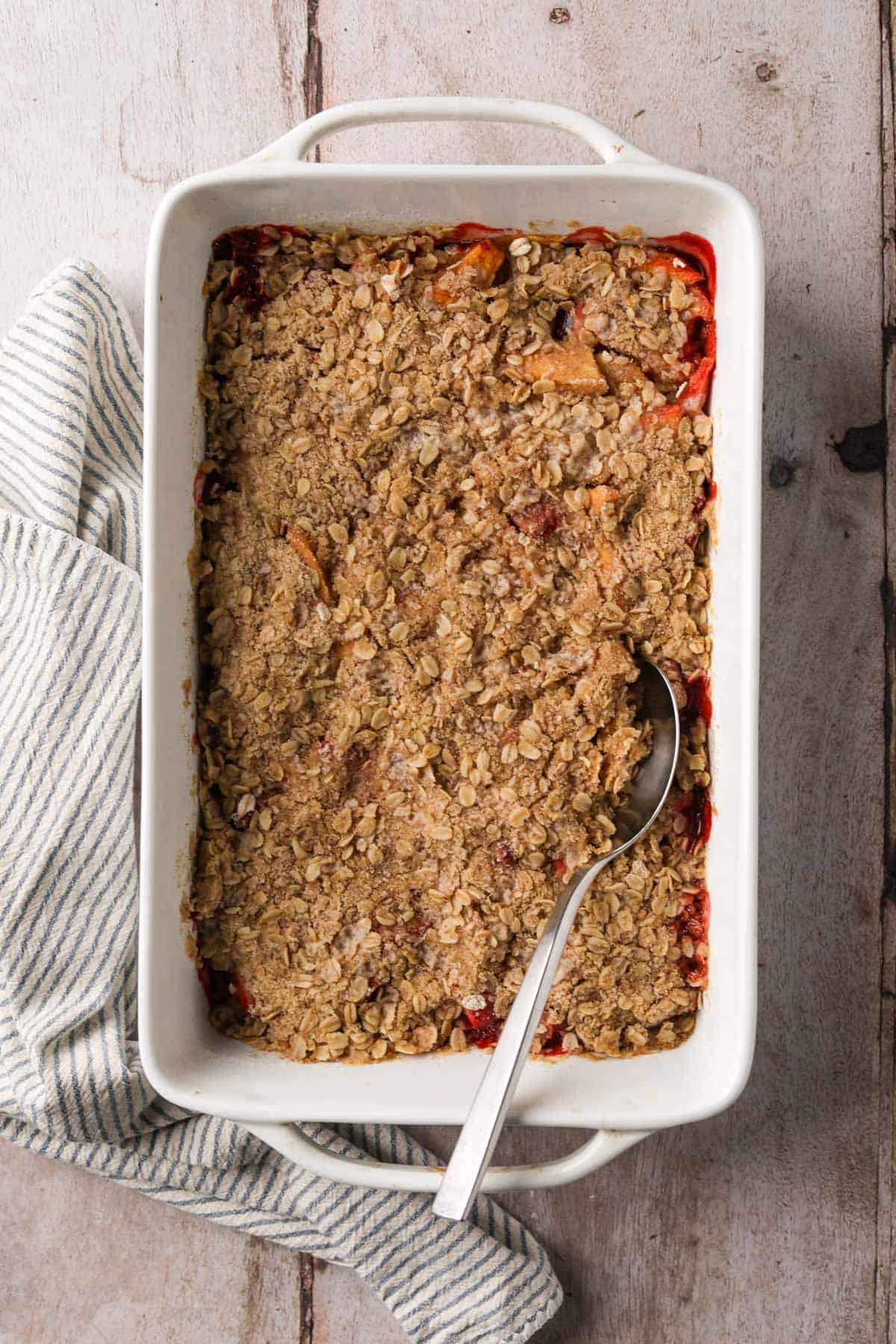 Baked strawberry peach crisp in a white ceramic 8x11 baking dish with a serving spoon.