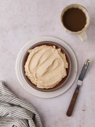 Overhead shot of coffee buttercream in a dish with a cup of coffee, offset spatula, and napkin to the side.