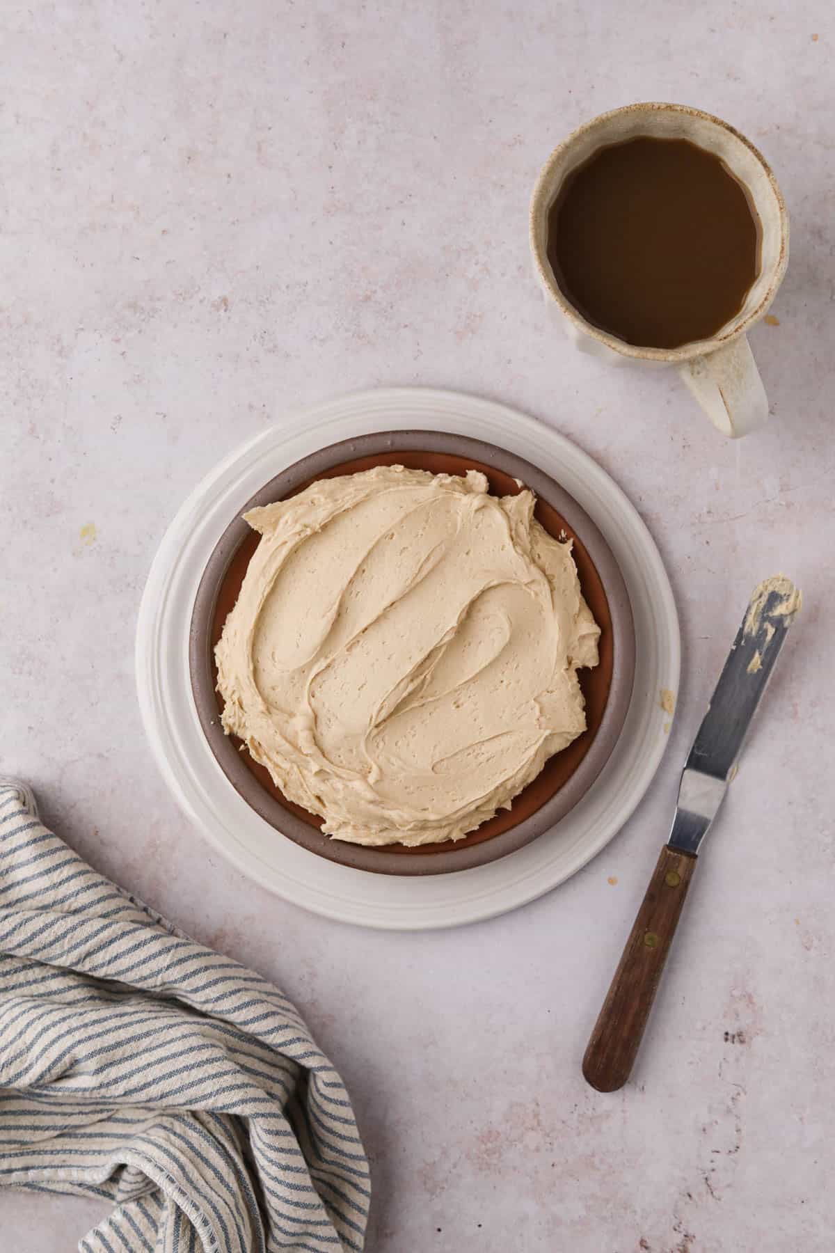 Overhead shot of coffee buttercream in a dish with a cup of coffee, offset spatula, and napkin to the side.