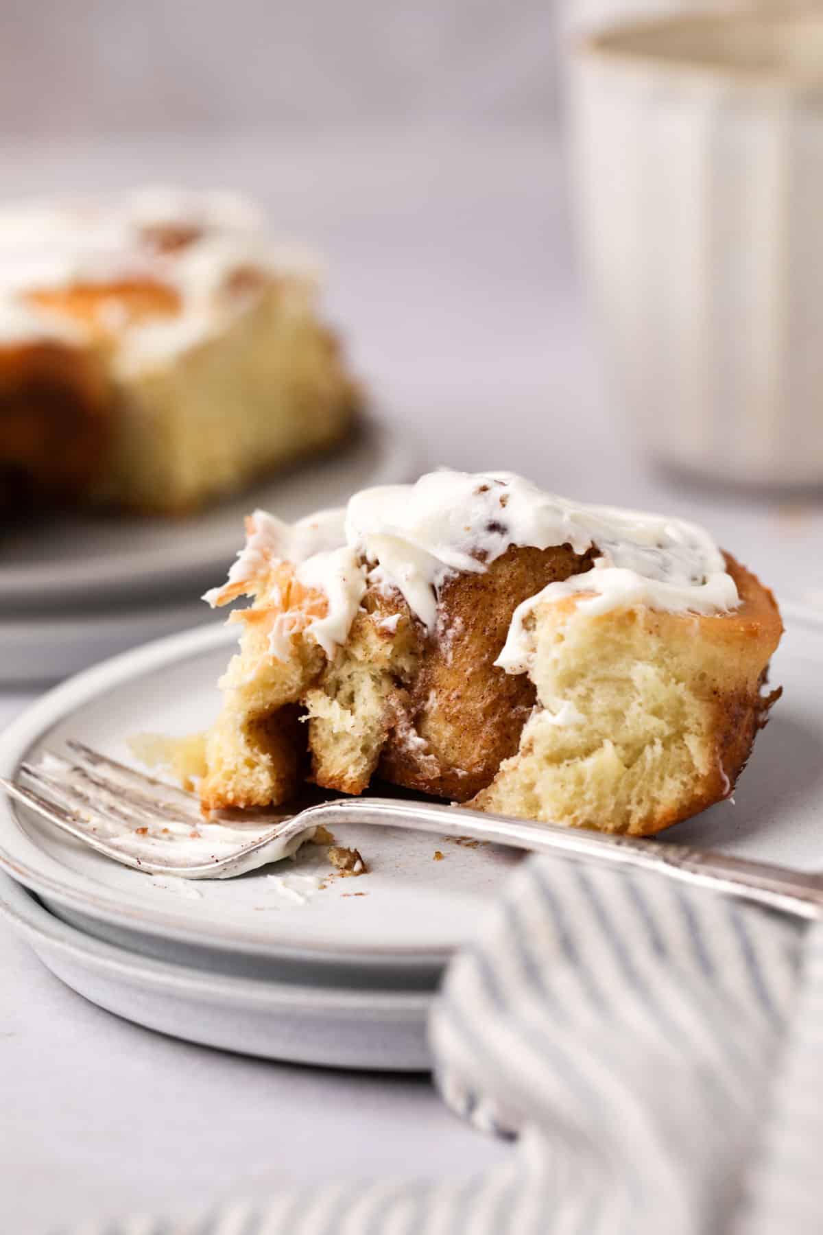 Close up of a homemade soft and fluffy cinnamon roll with cream cheese frosting set in plate with a fork and a bite taken out of it.
