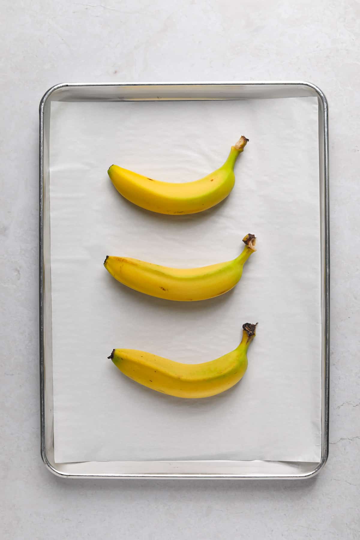 Three yellow bananas on a parchment paper lined sheet pan.