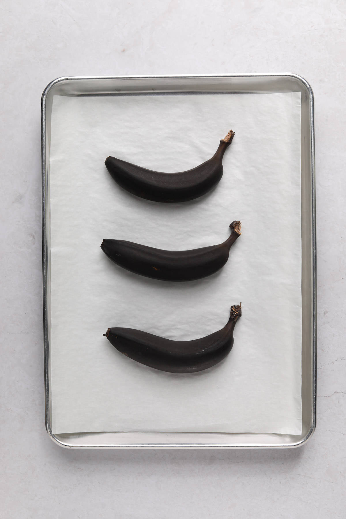 Three bananas on a parchment paper lined sheet pan that have been baked to black.