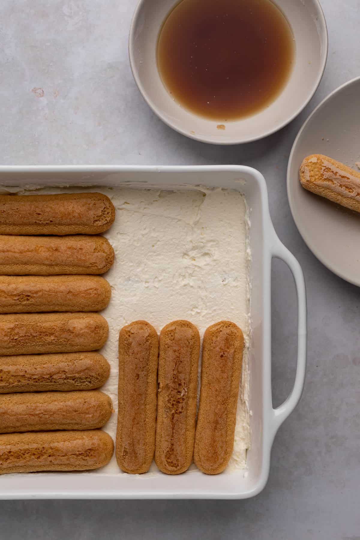 Ladyfingers dipped in espresso and arranges in a single layer of a white ceramic baking dish topped with cream filling and second layer oof ladyfingers.