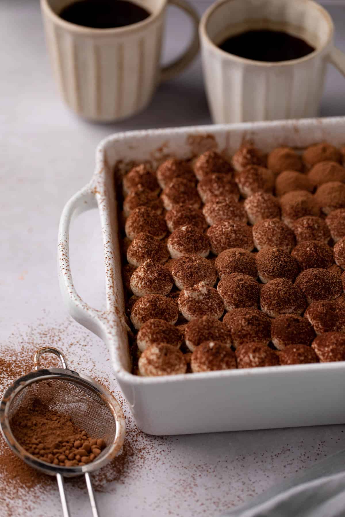 Angled shot of Easy tiramisu with piped creaming filling on top dusted with cocoa powder and coffee in the background.