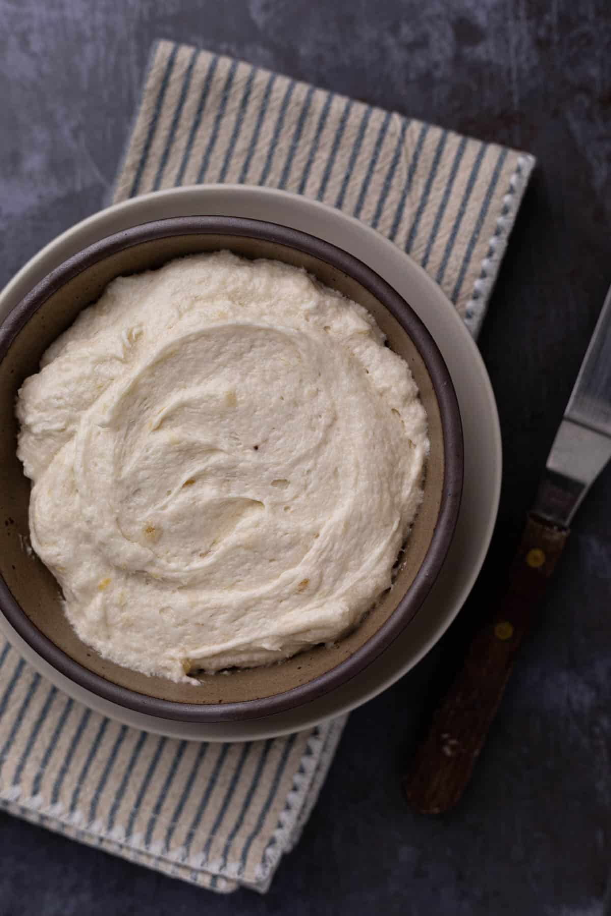 A bowl of banana buttercream frosting set on a flat surface with a striped napkin.