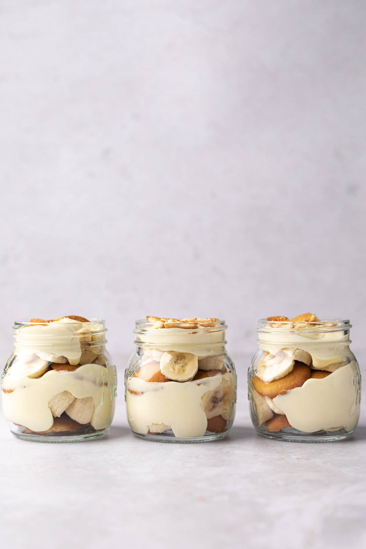 Three jars of banana pudding in a line topped with crushed vanilla wafers.