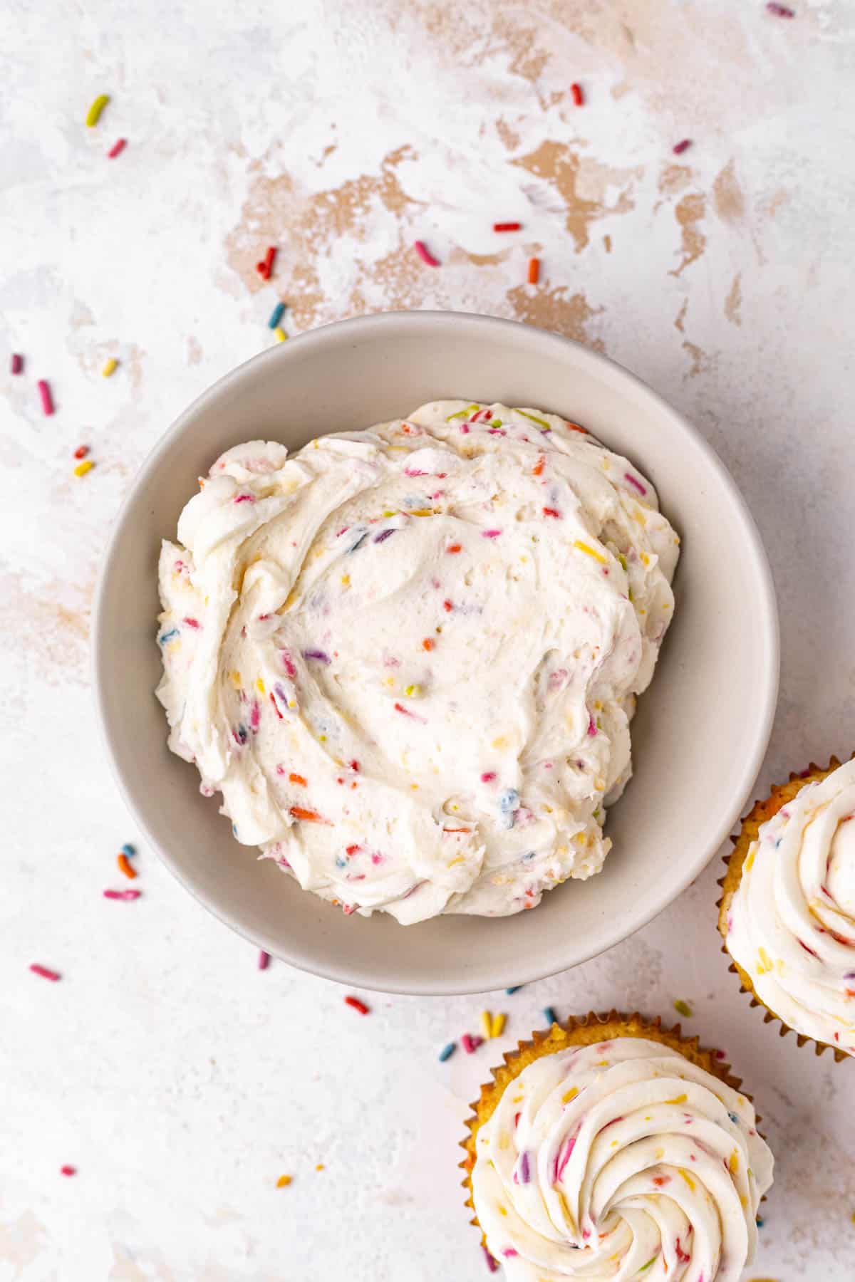 Overhead shot of a white bowl of funfetti buttercream frosting with two funfetti cupcakes set to the side.