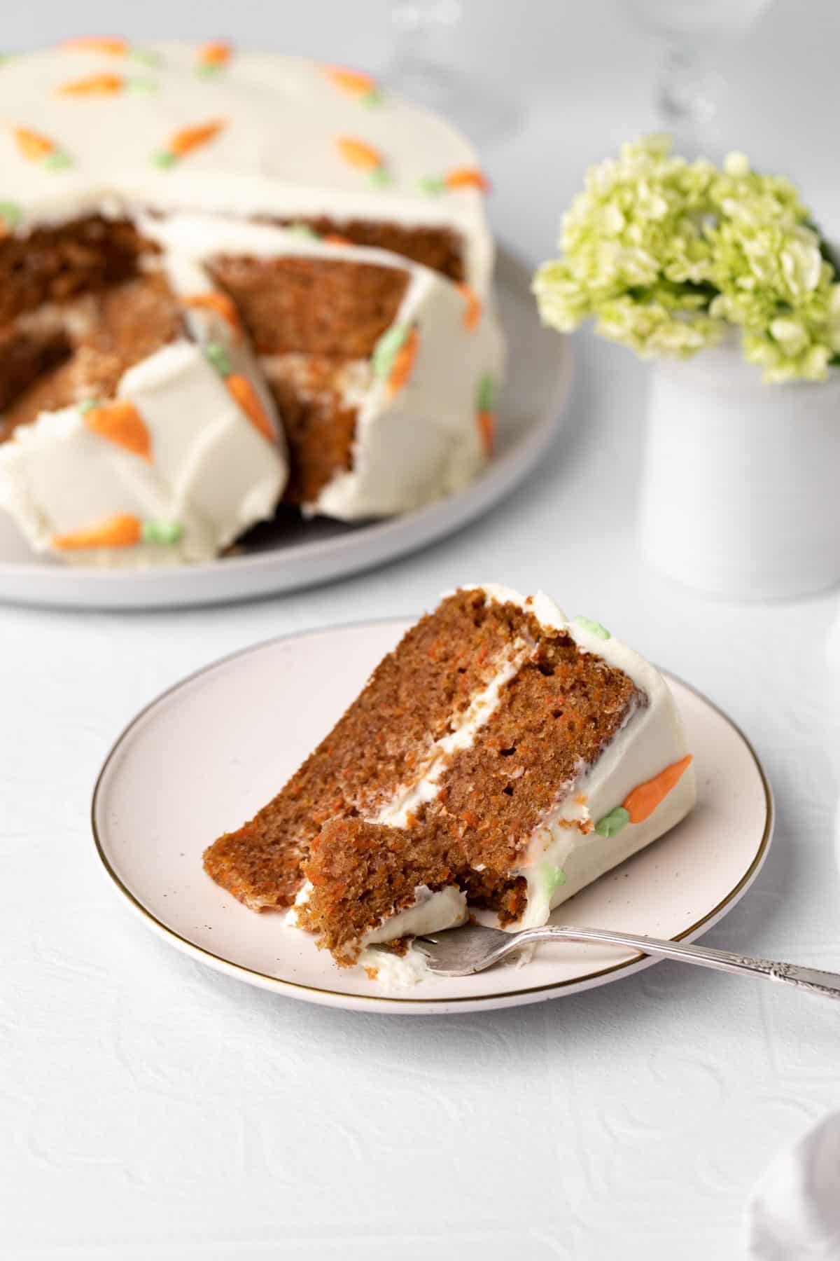 Slice of old fashioned carrot cake on a plate with a fork and the rest of the cake in the background.