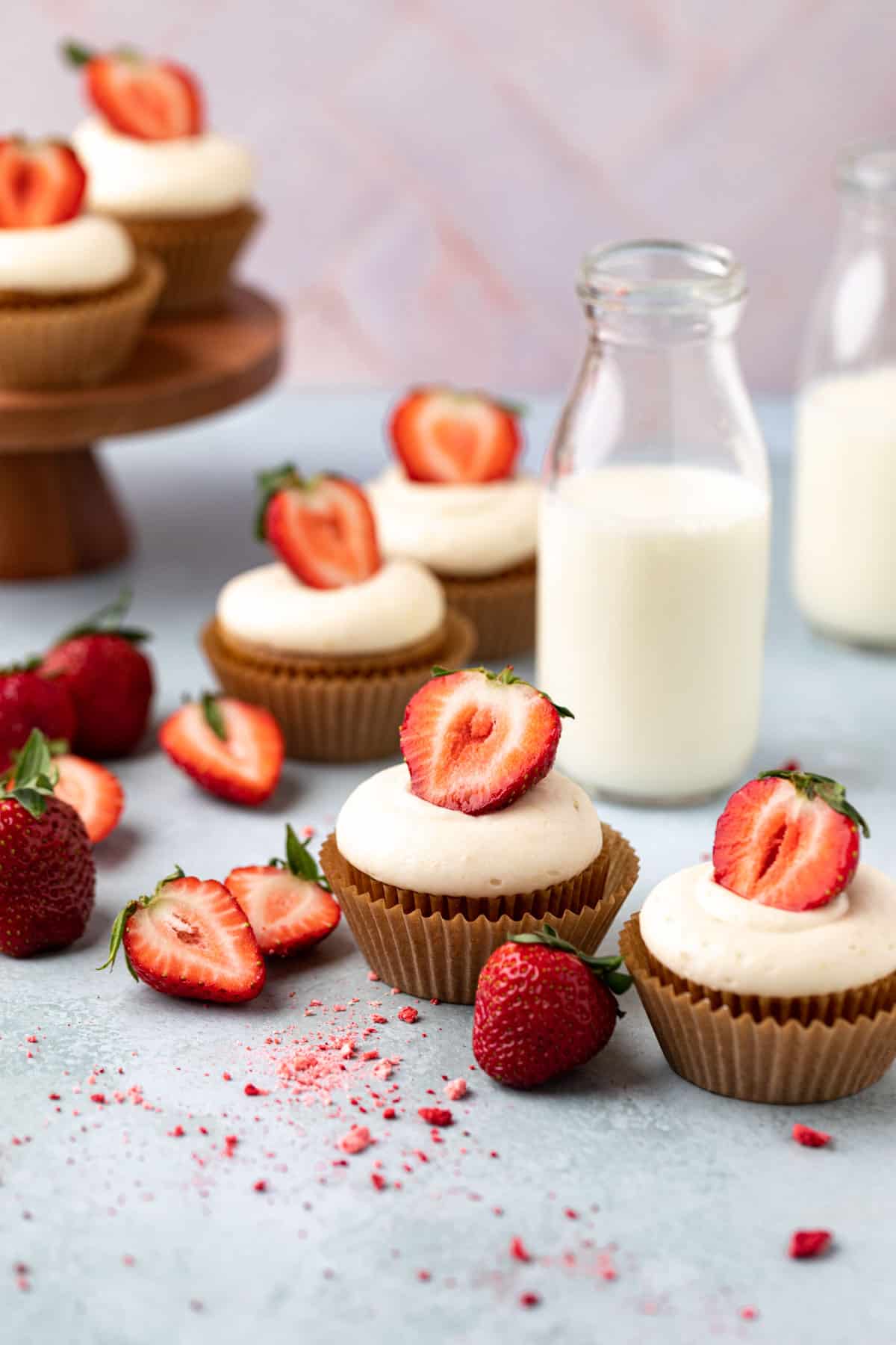 Fresh strawberry and banana cupcakes arranged on a flat surface with a small wood cake stand in the background and two glasses of milk.