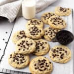 Chocolate chip sugar cookies set on a parchment lined black wire cooling rack with a cup of mini chocolate chips and glass of milk.