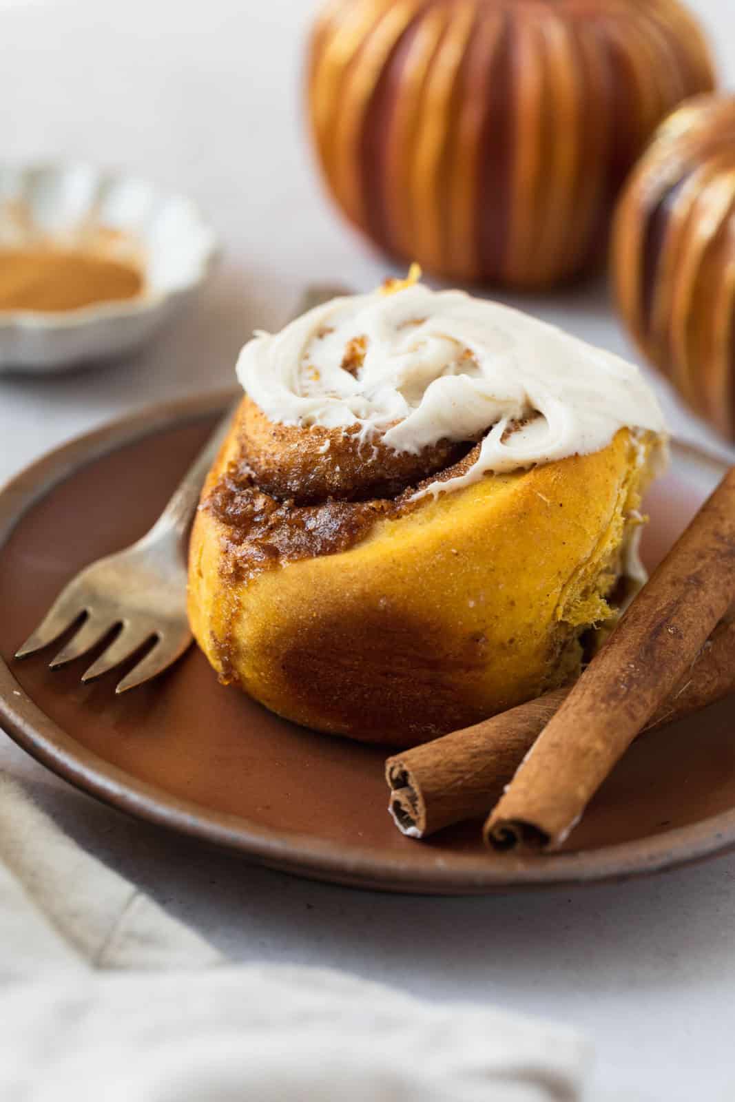 Close up of a pumpkin cinnamon roll in a plate with cinnamon sticks.