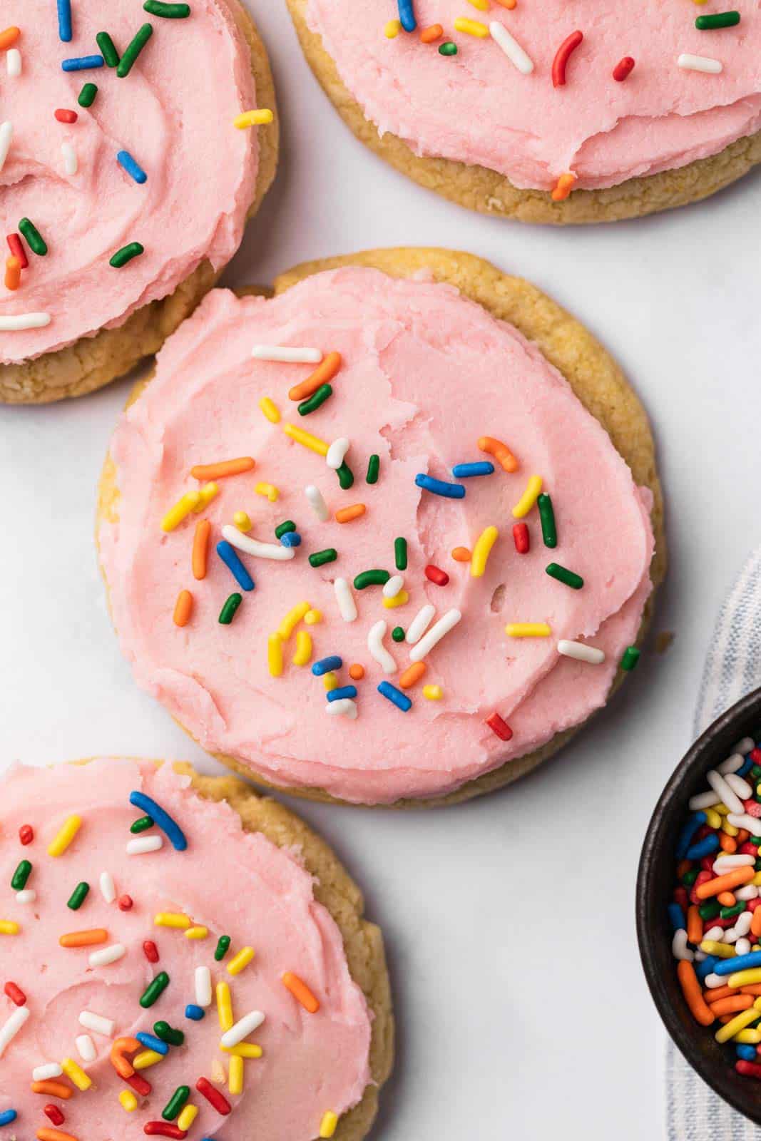 Close up of soft vanilla sugar cookies frosted with pink buttercream and topped with rainbow sprinkles.