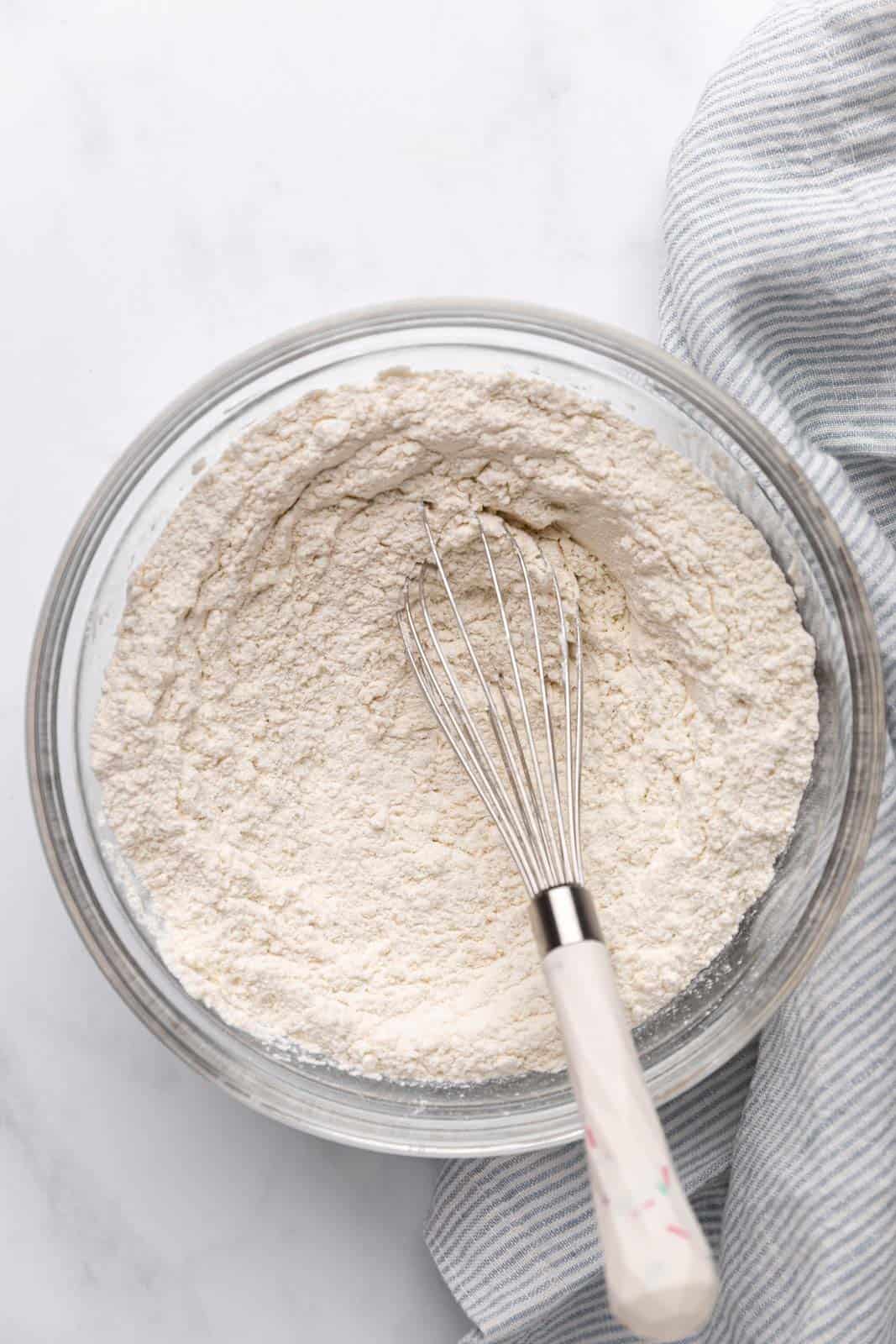 Whisked dry ingredients for sugar cookies in a glass mixing bowl set on a flat surface with a whisk in the bowl.