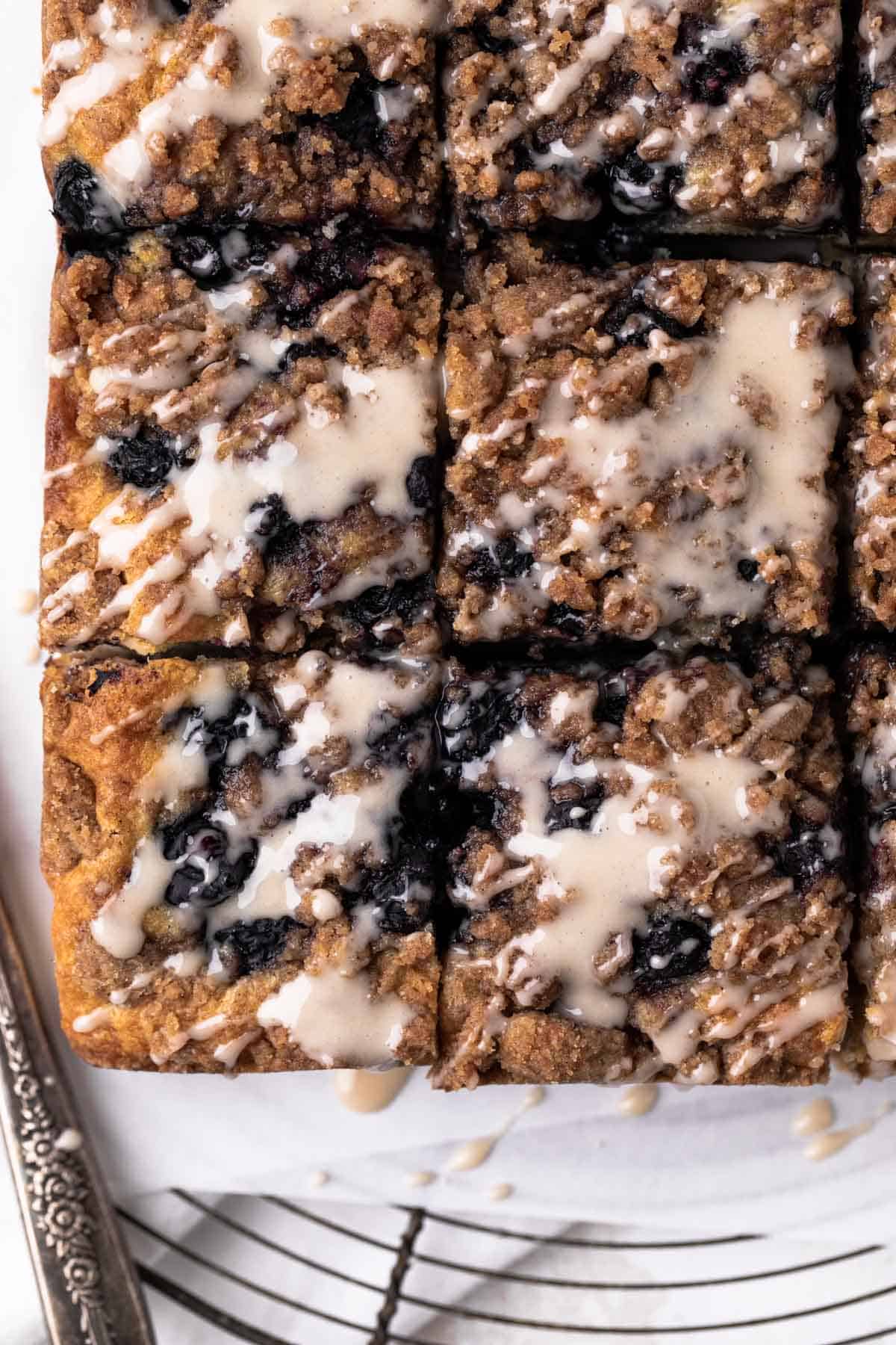 Vanilla glaze drizzled over squares of blueberry coffee cake set on a parchment lined round wire rack.