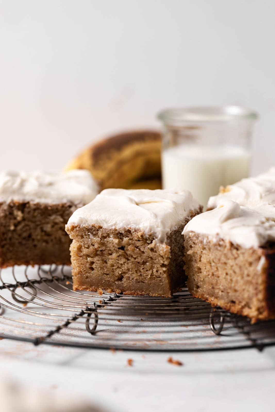 Close up of squares of banana snack cake with brown sugar cream cheese frosting spread on top and set on a round wire rack.