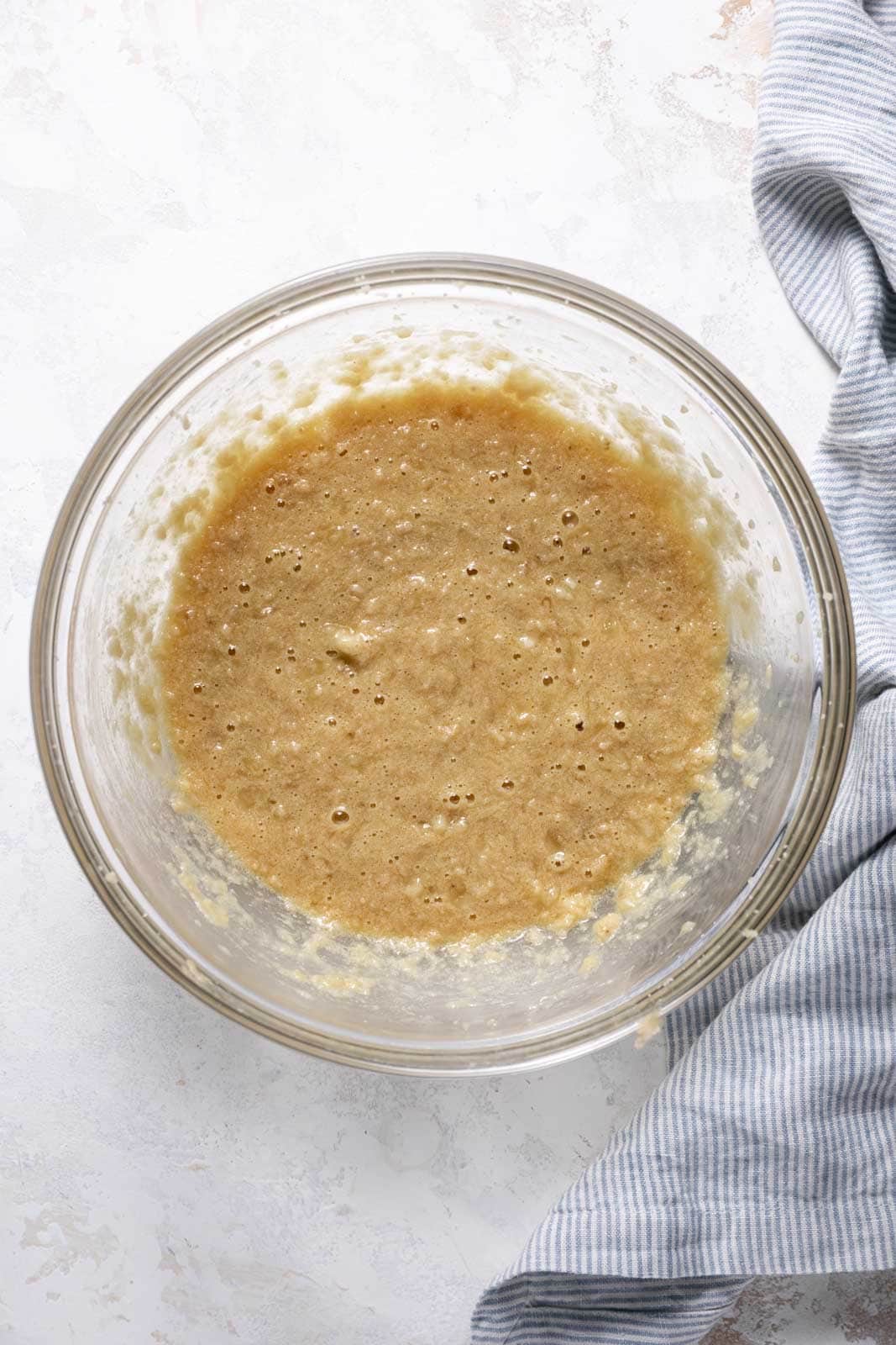 Mashed banana, eggs and vanilla mixed into creamed butter and sugar in a glass mixing bowl set on a flat surface.