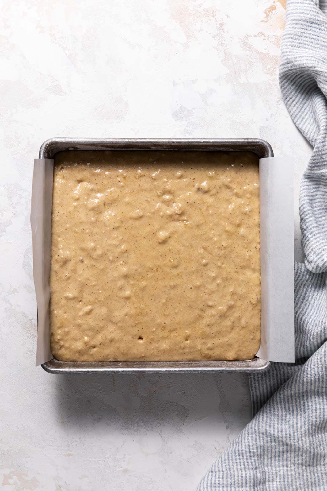 Banana snack cake batter in a metal square pan lined with parchment paper set on a flat surface.