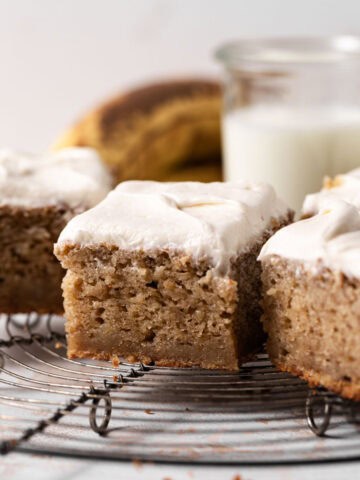 Close up of squares of banana snack cake with brown sugar cream cheese frosting spread on top and set on a round wire rack.