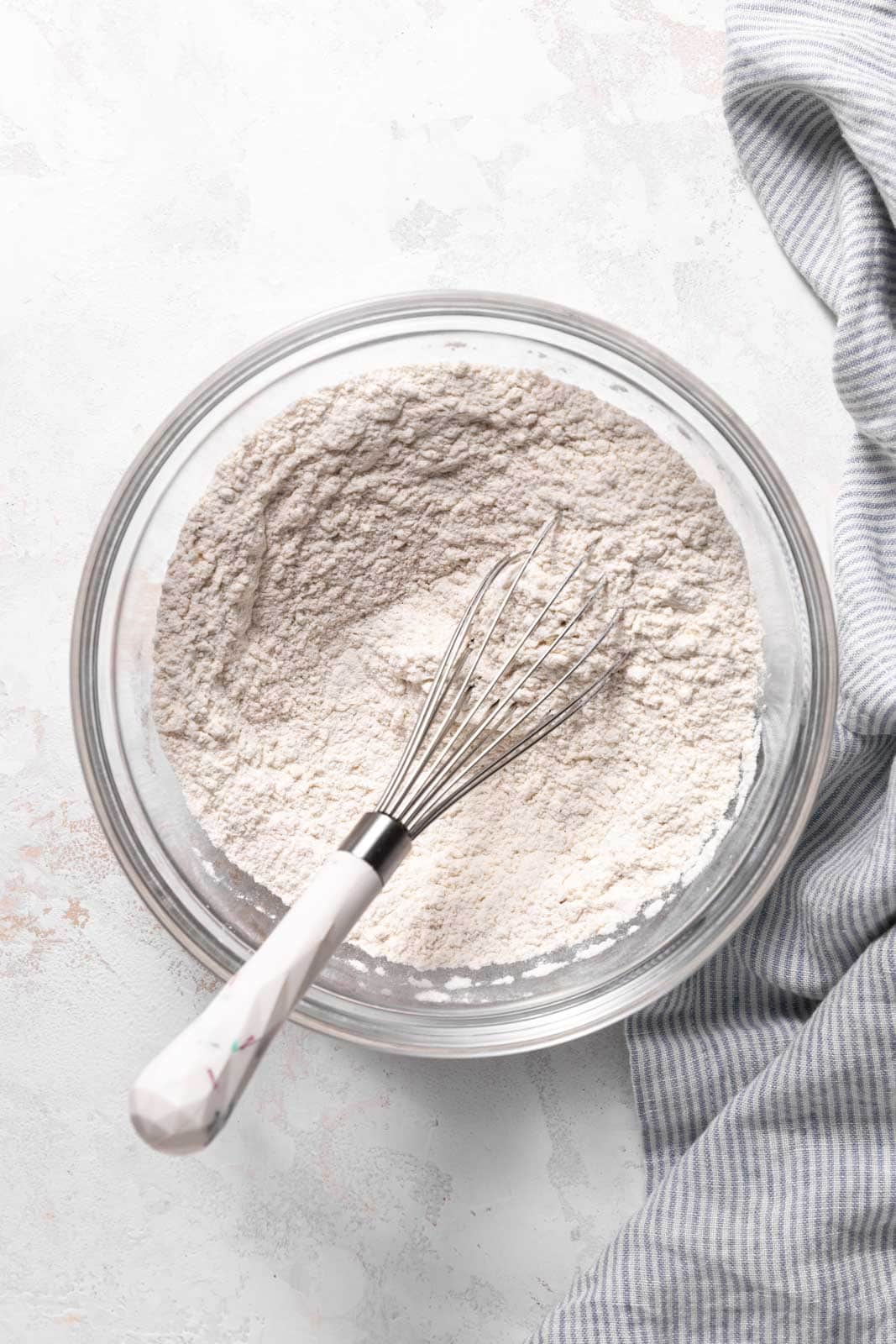Whisked dry ingredients in glass mixing bowl with a whisk set on a flat surface.