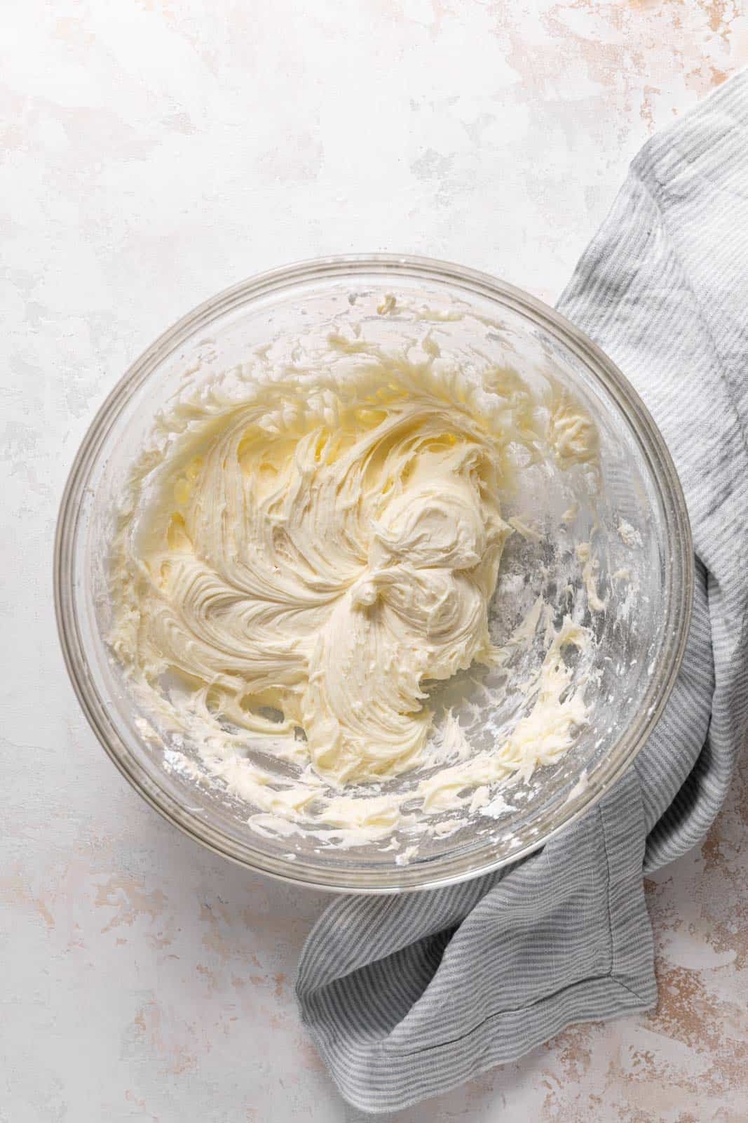 Powdered sugar mixed with creamed butter and cream cheese in a glass mixing bowl set on a flat surface.